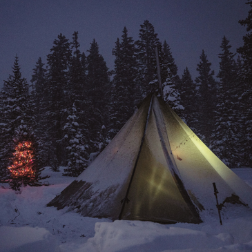 Snow camp with 12-person tipi glowing from inside, stove pipe smoking at peak