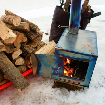 a fire glow inside an XL titanium wood stove with stove pipe, split firewood piled nearby 