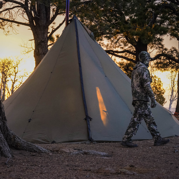 hunter walks past a 12 person tent in fading light, stove pipe coming through stove jack visible against sunset glow
