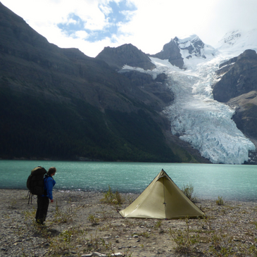 backpacker stands next to Cimarron tent in high mountain meadow, glacier and glacial lake in the background