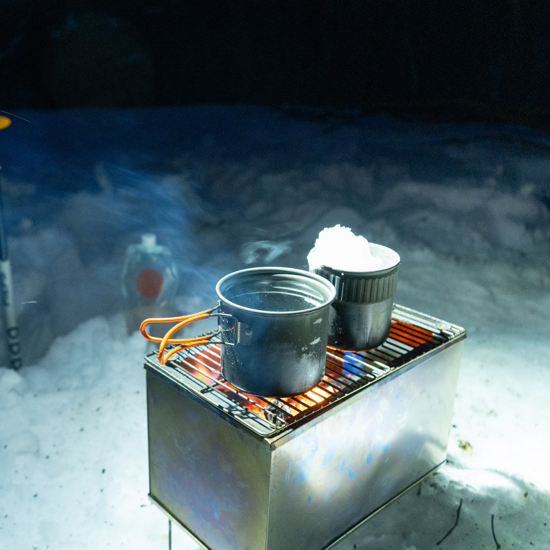 Backcountry stove with grill top, glowing embers and visible flames, heating a pot for hot food.