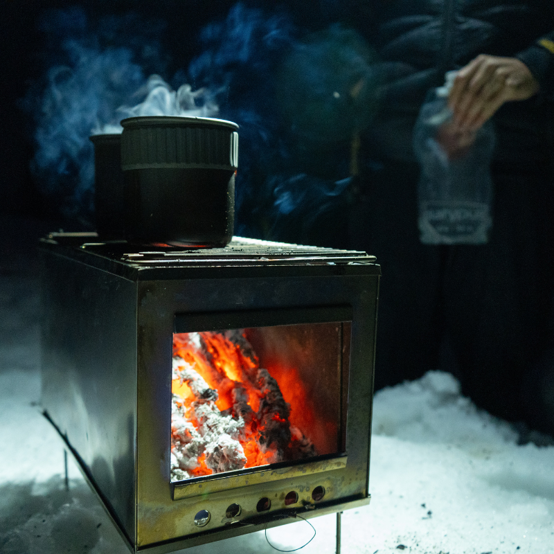 Flames visible through glass door of Seek Outside stove with grill top, boiling a pot of water.