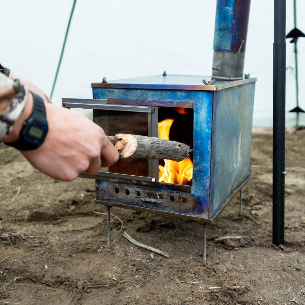person adds wood to the fire inside a seek outside titanium stove, fire visible through the Glass Door