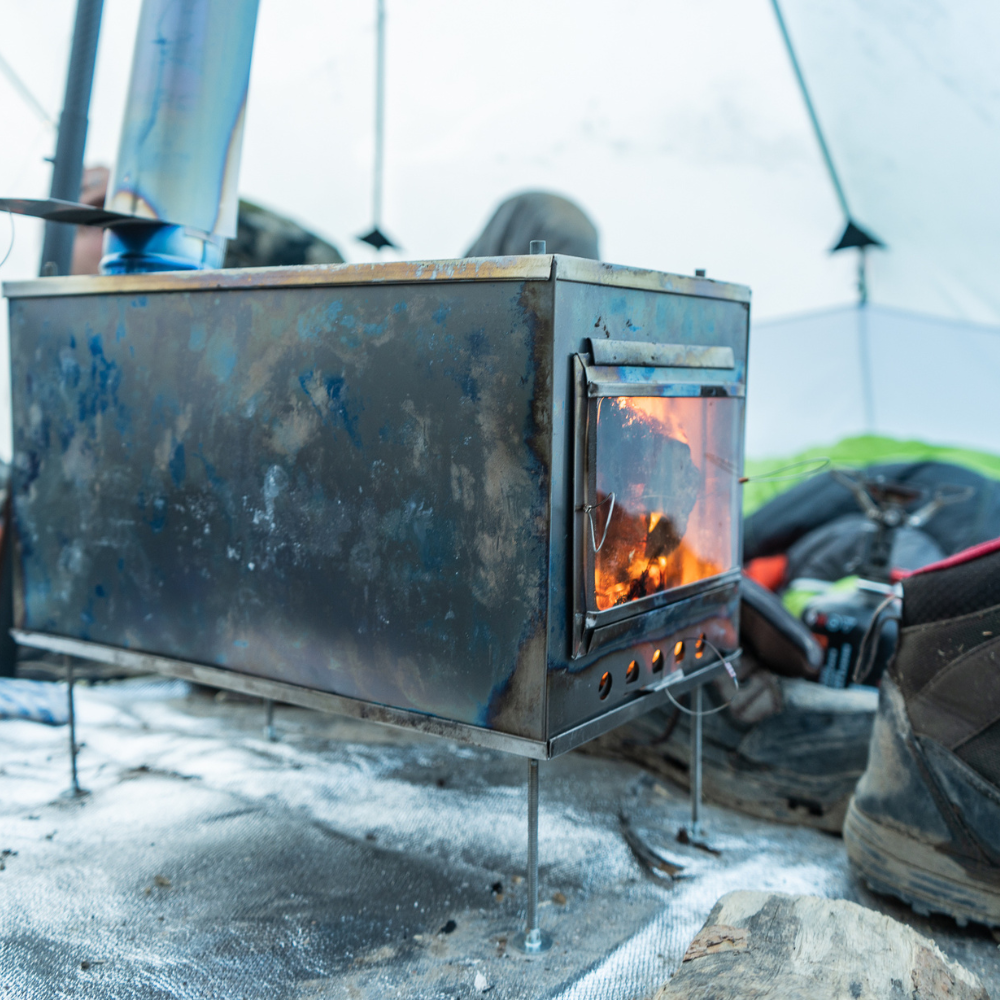 Tent stove with glass door showing bright orange flames and glowing embers inside the firebox