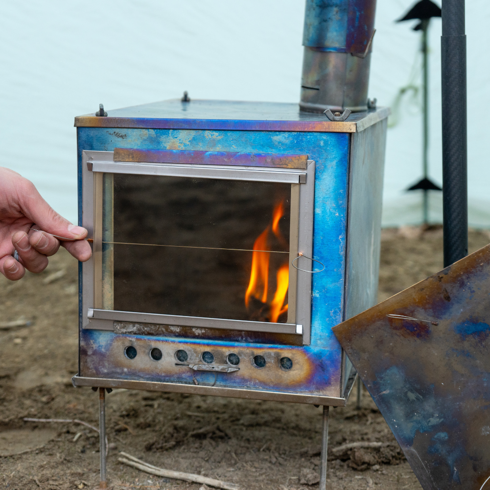Glass door on a Seek Outside tent stove glowing from the fire inside, providing a clear view of the flame and lighting the surrounding area