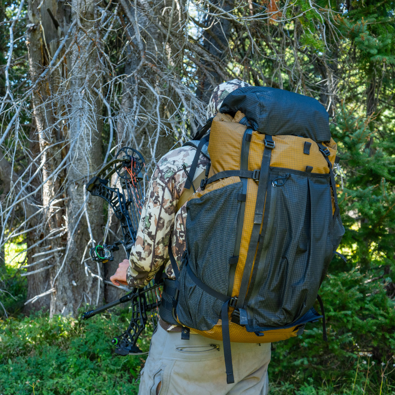 a man holding a hunting crossbow waring a goshawk backpack in a dense forest
