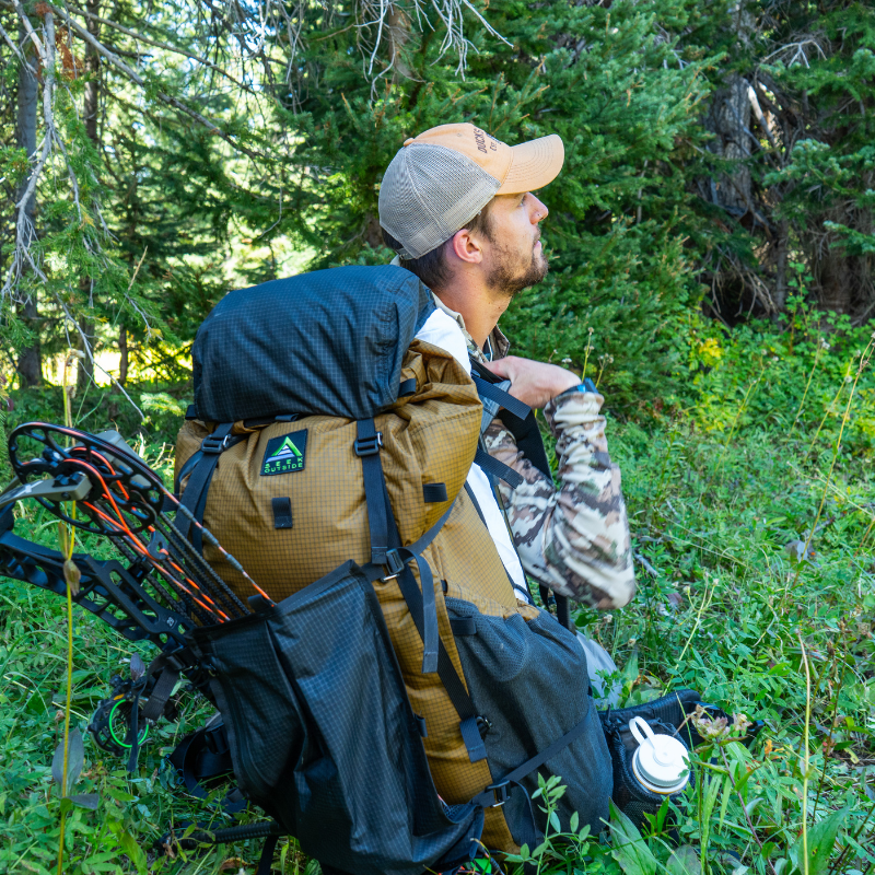 a man sits on the ground with a seek outside goshawk backpack. the detachable talon on the backpack is used to attach a crossbow to the backpack
