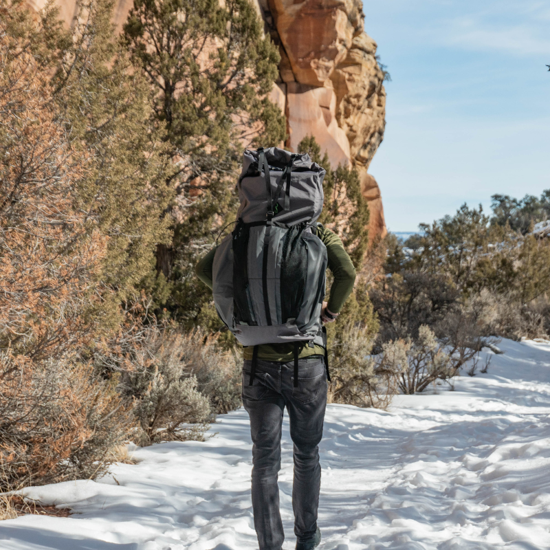 a man wearing a Gila backpack stands on a snowy trail with rocky cliffs in the background