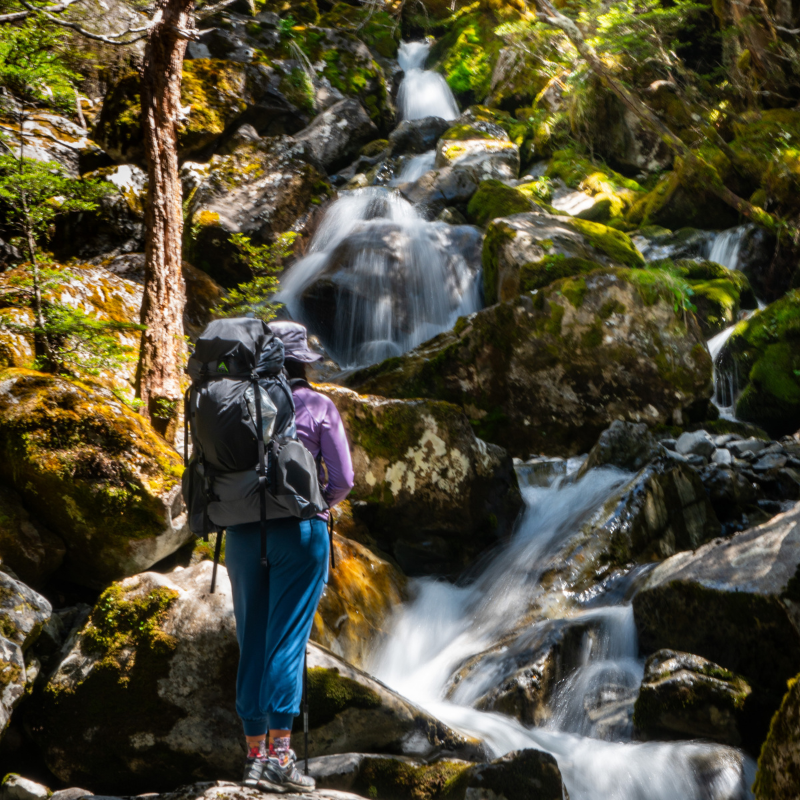 a woman wearing a unaweep backpack stands at the bottom of a cascading mountain waterfall