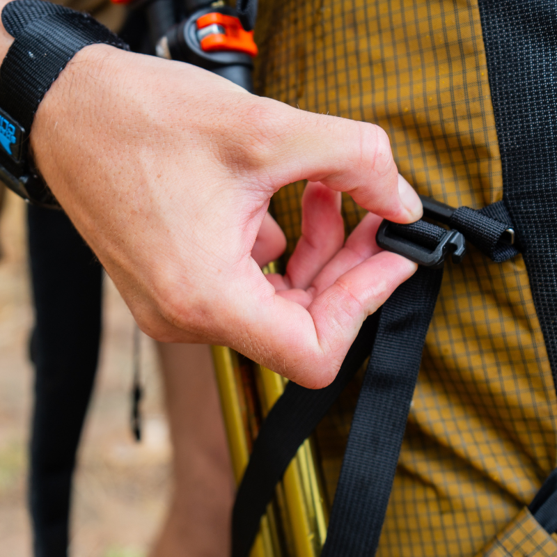 close up view of a hand hooking a gatekeeper strap to a loop on a Coyote brown backpack to secure trekking poles in the backpack side pocket