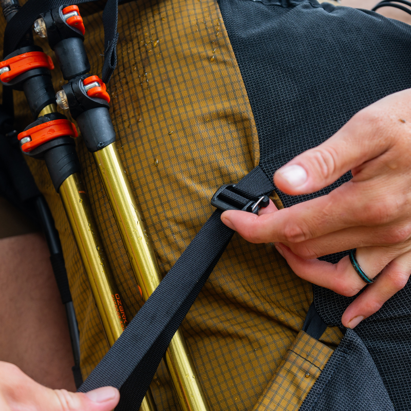 close up view of a hand hooking a gatekeeper strap to a loop on a Coyote brown backpack to secure trekking poles in the backpack side pocket