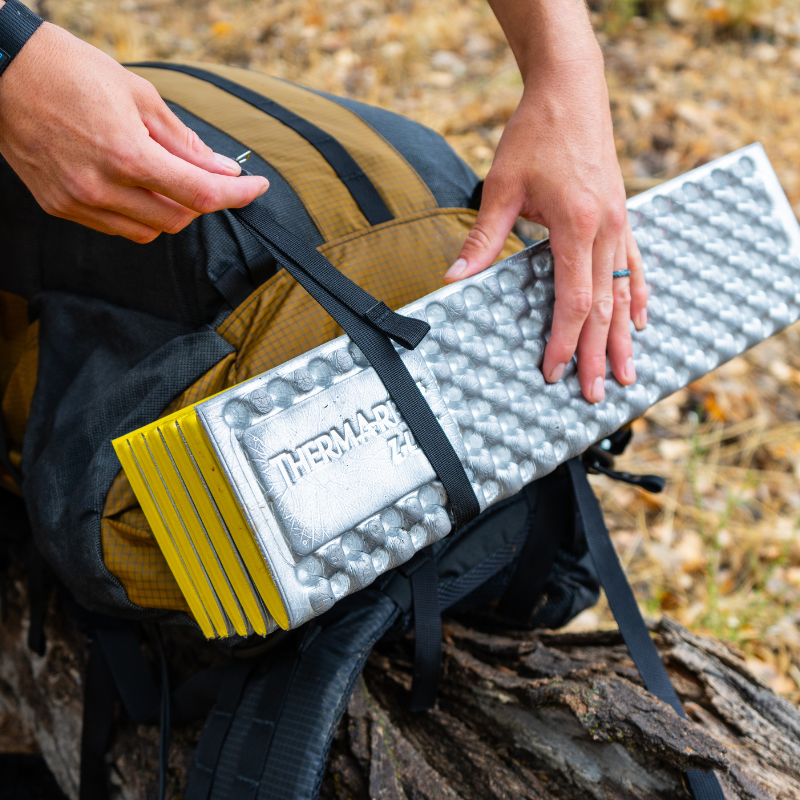 close up view of a hand hooking a gatekeeper strap to a loop on a Coyote brown backpack to secure a sleeping pad to the bottom of a backpack