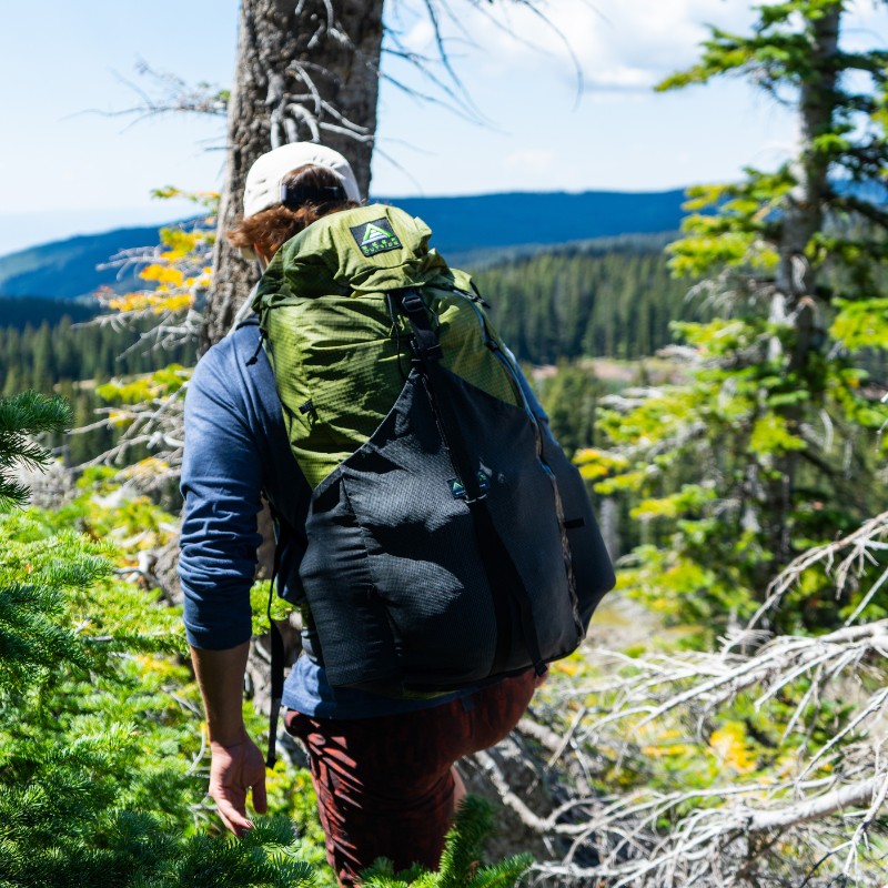 Hiker emerging from dense green forestry trail, Silverton pack blending with earthy tones, front stretch pockets filled with gear