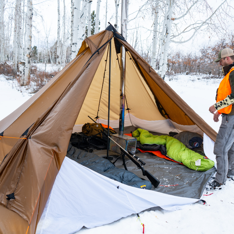 a man stands in the snow next to a redcliff tent, sleeping bags, a titanium stove and camping gear inside the tent