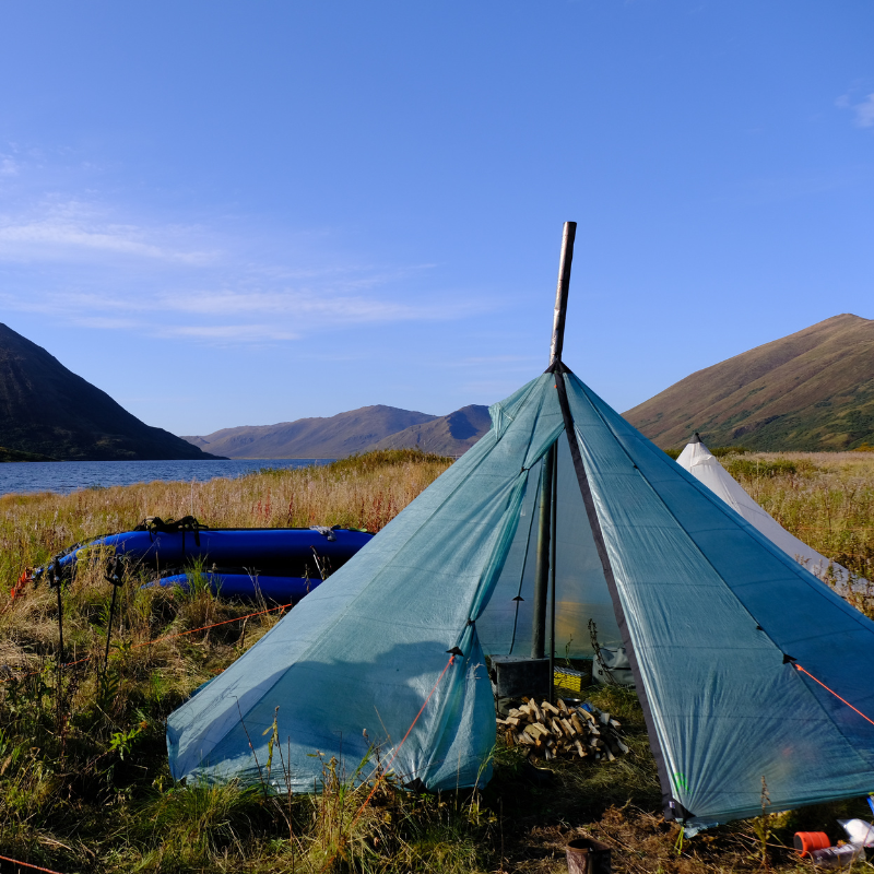Sage Dyneema Redcliff tent sits on a grassy riverbank, inside the tent is a wood stove, chopped wood and camping gear