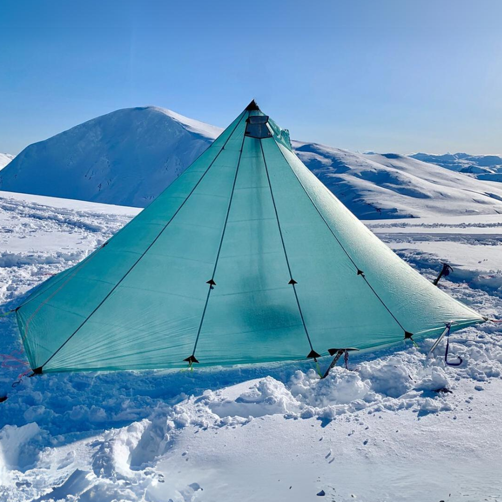 a dyneema redcliff tent is set up in a snowy hillside, snow covered mountains and clear blue skies in the background