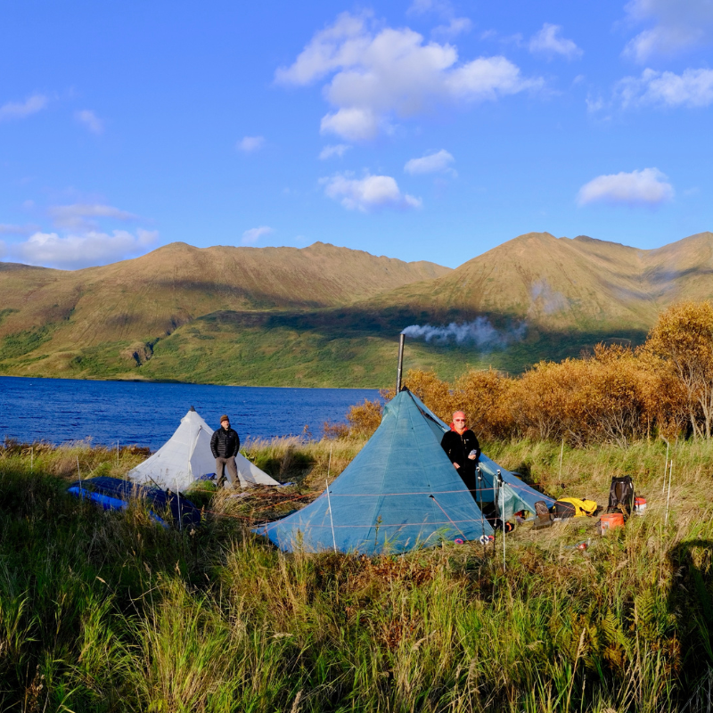 A Redcliff Dyneema shelter sits in a grassy clearing at the side of a lake, hills and blue skies in the background