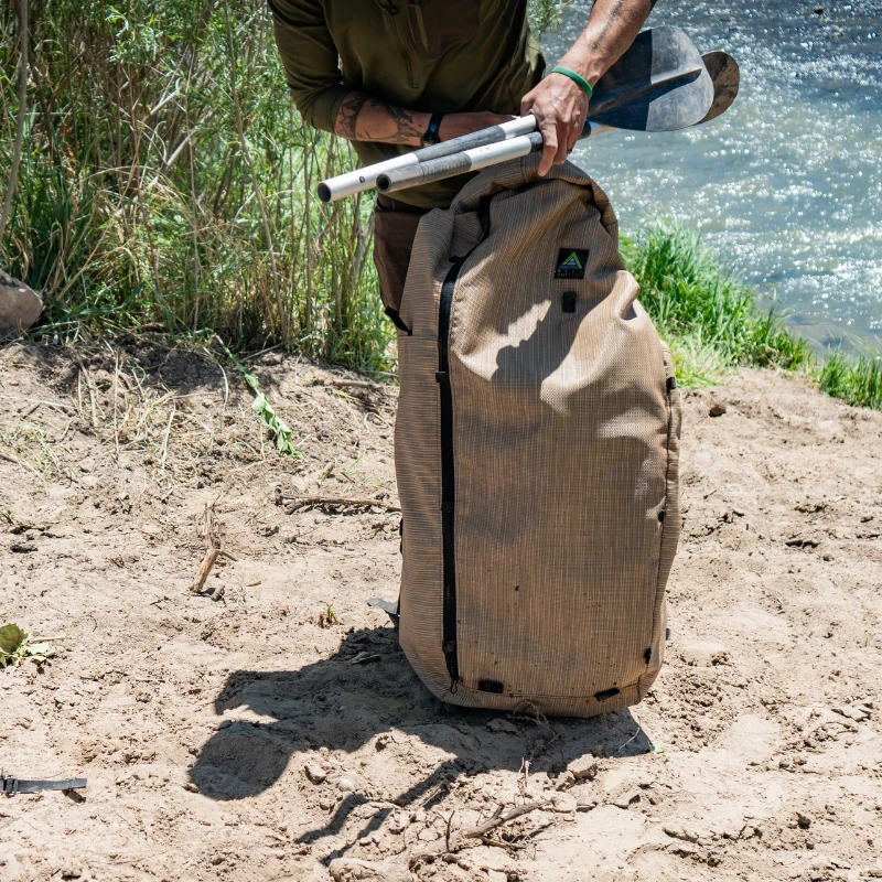 a wetlands backpack sits in the sand on a riverbank, a man adjusts the contents of the backpack to accommodate two raft oars