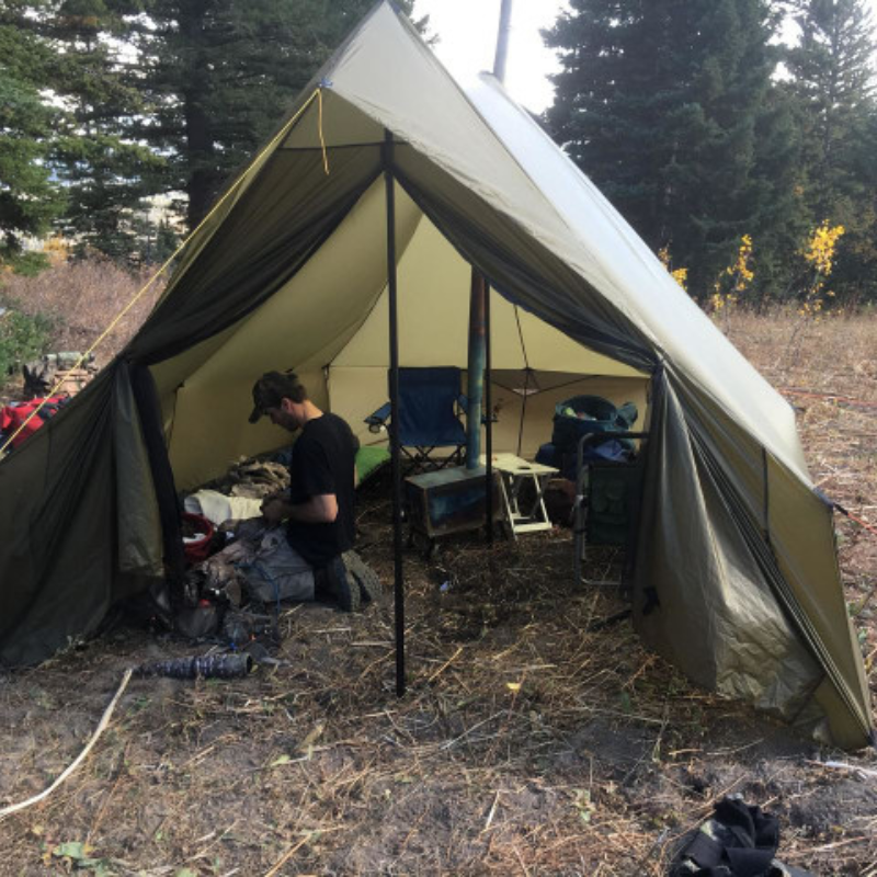 a man kneels inside a spacious Courthouse tent surrounded by camping gear and chairs, in a forested area