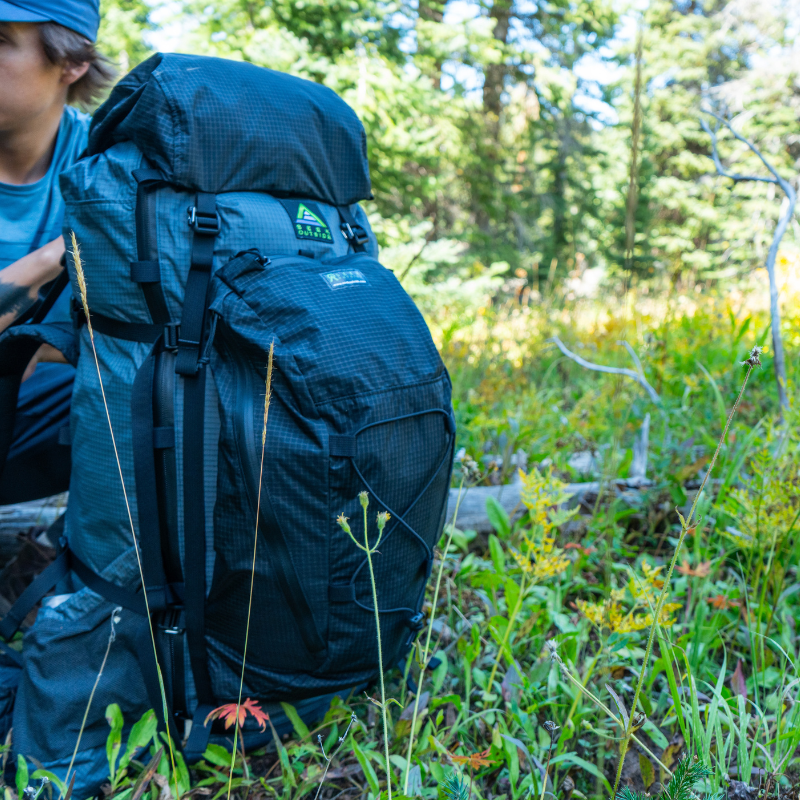 black merlin backpack connected to a gray backpack, backpack is on the ground in a grassy field with a man holding it
