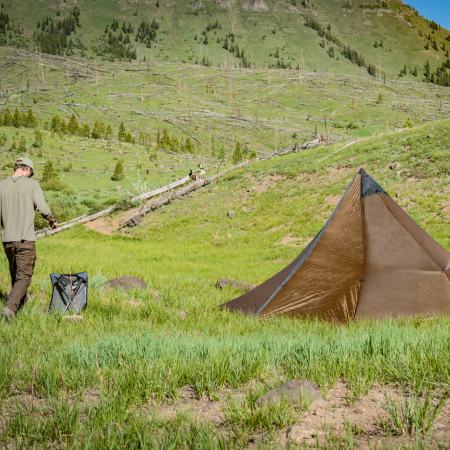 backpacker stands near a sitting saddle set up in a grassy valley, a brown tent is set up nearby