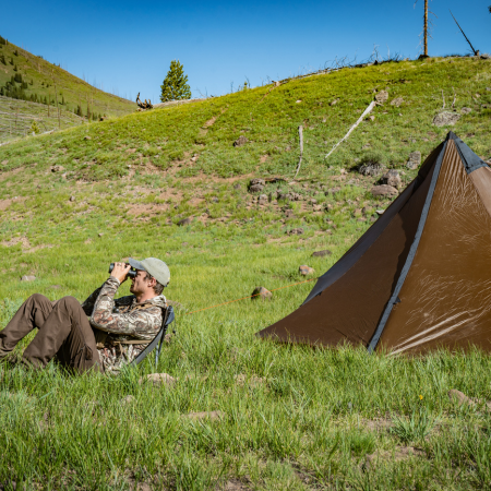 a man sits in a sitting saddle and looks through binoculars, a tent is set up behind him in the grassy valley