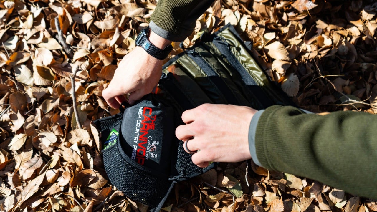 a man puts a game bag into the mesh pocket of a flat roll, the flat roll lays on the leaf covered ground