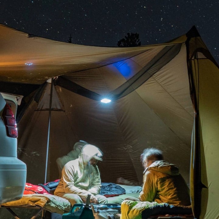 tow men sit on cots inside a bookcliff tent, the nightime scene is illuminated by a light inside the bookcliff attic hanging from the middle of the tent