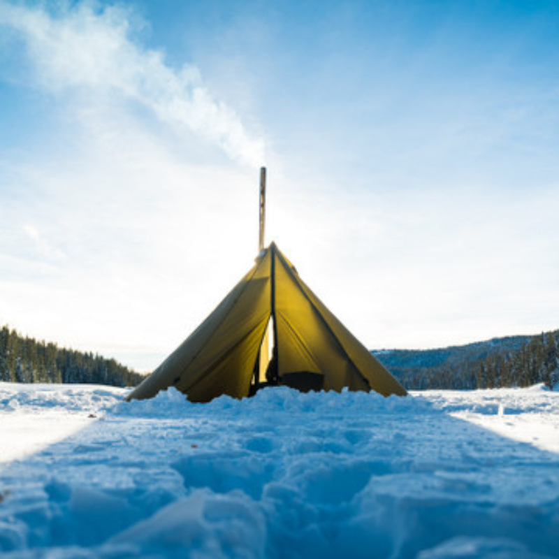 Seek Outside teepee pitched on compact snow with twisted stakes holding guy lines, bright sunlight casting sharp shadows on pristine snow 