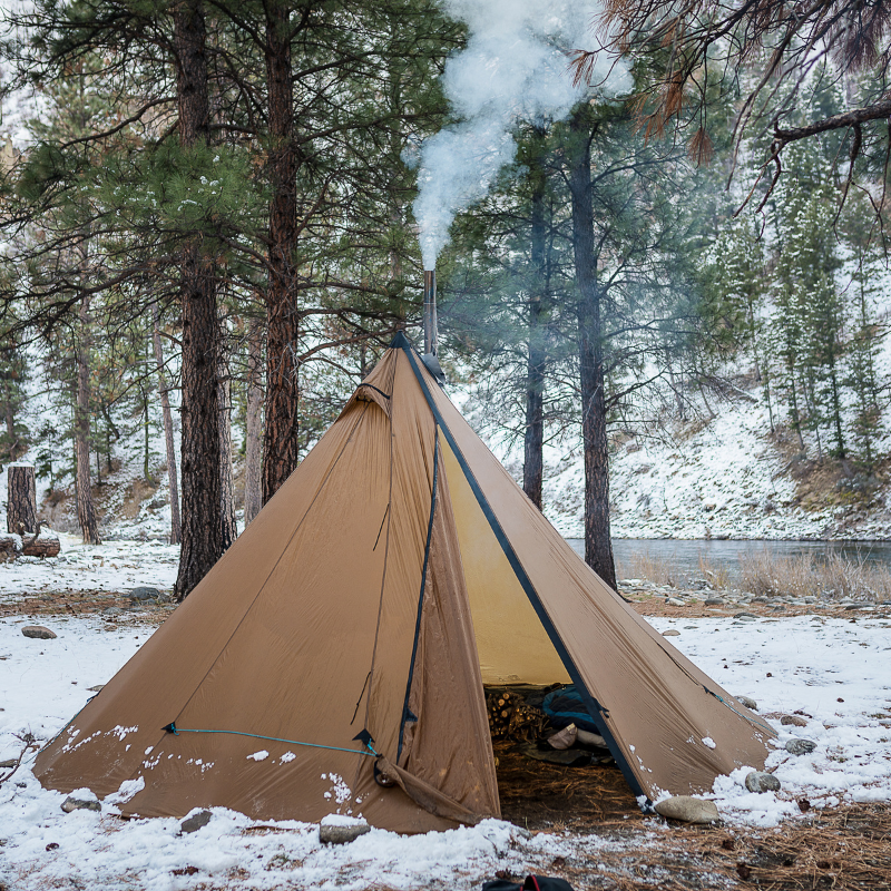 8 person tipi pitched in a snowy forest, stove, firewood and gear visible through open door