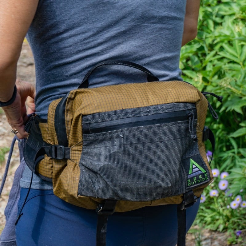 Mesa hip pack in coyote color being worn by a woman next to green bushes