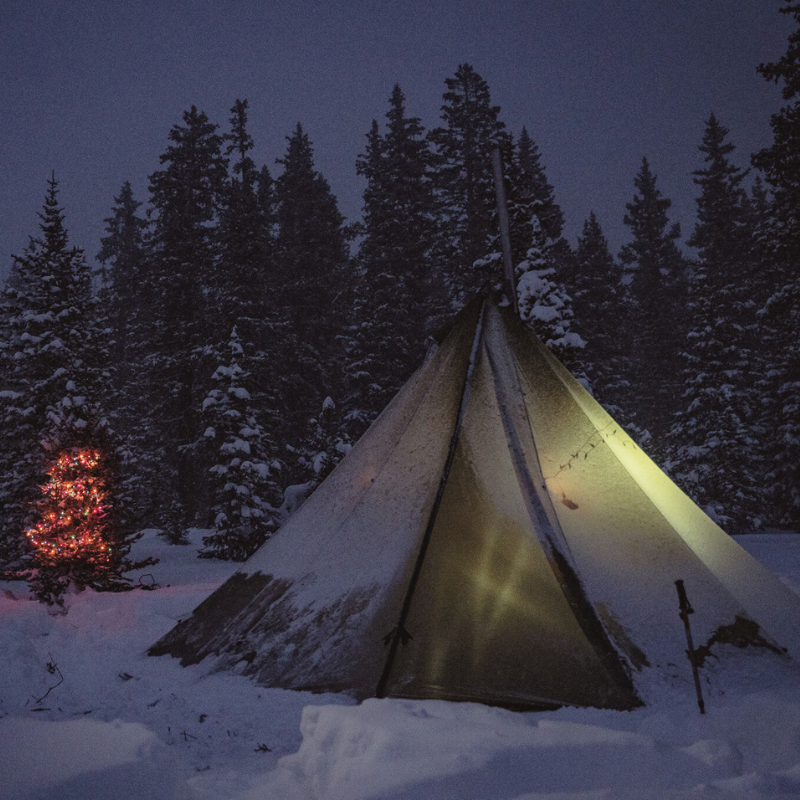 Snow camp with 12-person tipi glowing from inside, stove pipe smoking at peak