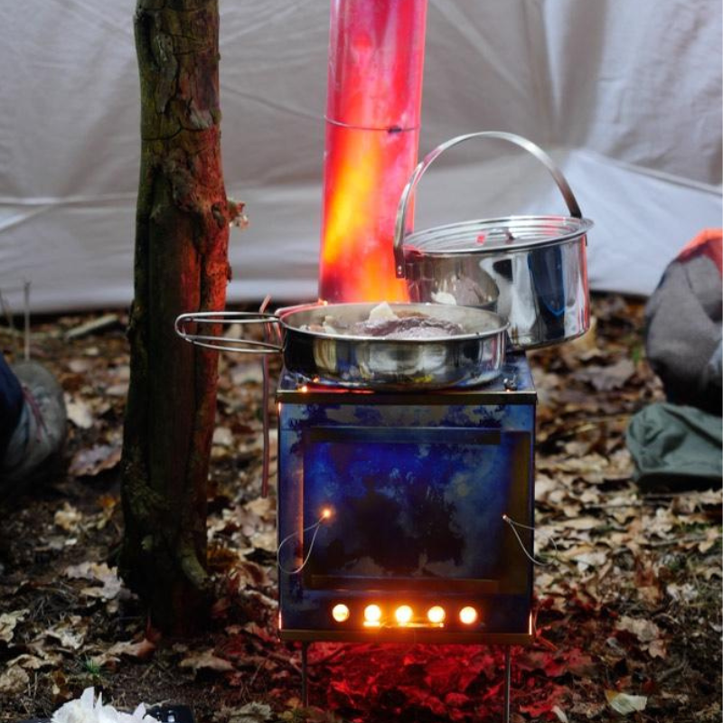 titanium stove with fire inside set up inside a tent, stove pipe burning red hot, flames visible through from dampers, cooking pot and skillet with steak cooking on top of stove 