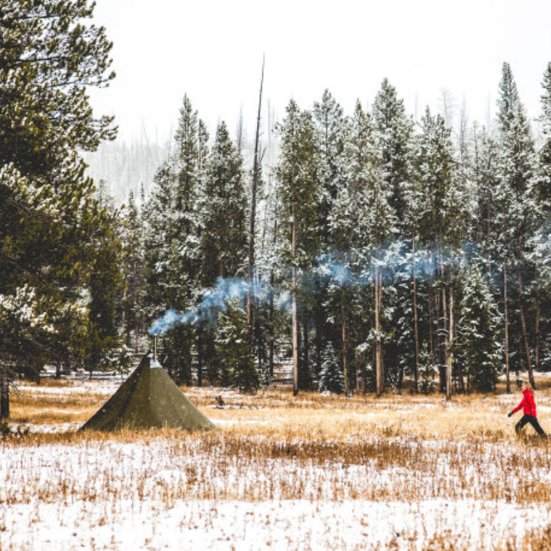 Mountain backcountry camp with 6-person tipi shelter, stove smoke rising, and snowy pine forest in background