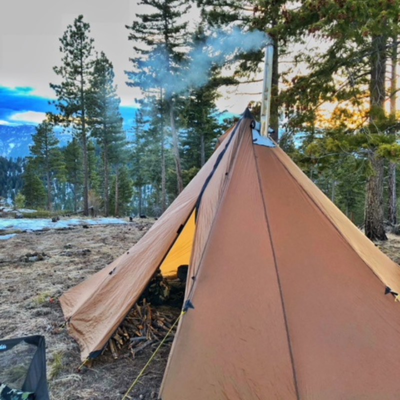 Hunting basecamp inside Redcliff tent with titanium stove, split firewood, and gear stowed on one side leaving 3-person sleeping area