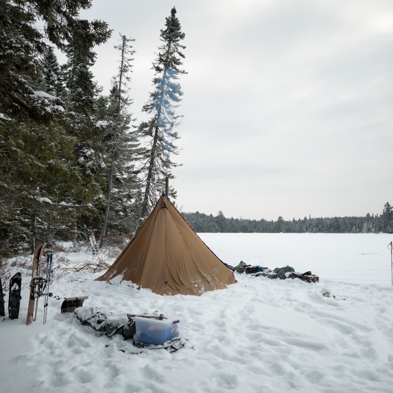 Tipi tent with snow piled around sod skirt for insulation, stove pipe emitting faint smoke in clear winter air