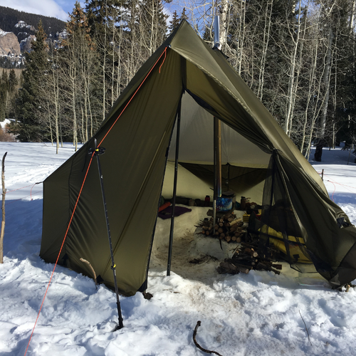 Courthouse tent set up in a snowy field, the open doors show a camp stove with wood piled nearby and camping gear inside the tent