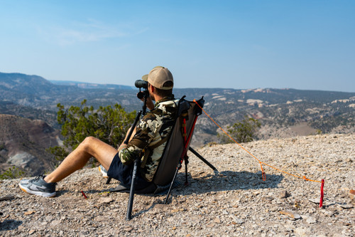 hunter sits in sitting saddle and looks through binoculars on a rocky hilltop