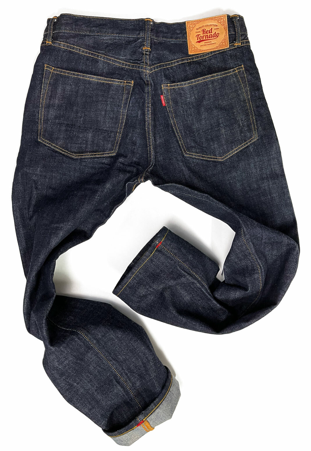 The back view of the Red Tornado selvedge denim jeans features well-defined contrast stitching along the pockets and seams. The jeans are artistically shaped to emphasize the chain stitch hemming the turned-up cuff, with a small red bartack detail visible on the hem. A leather brand patch on the waistband adds a finishing touch to the garment's dark denim.