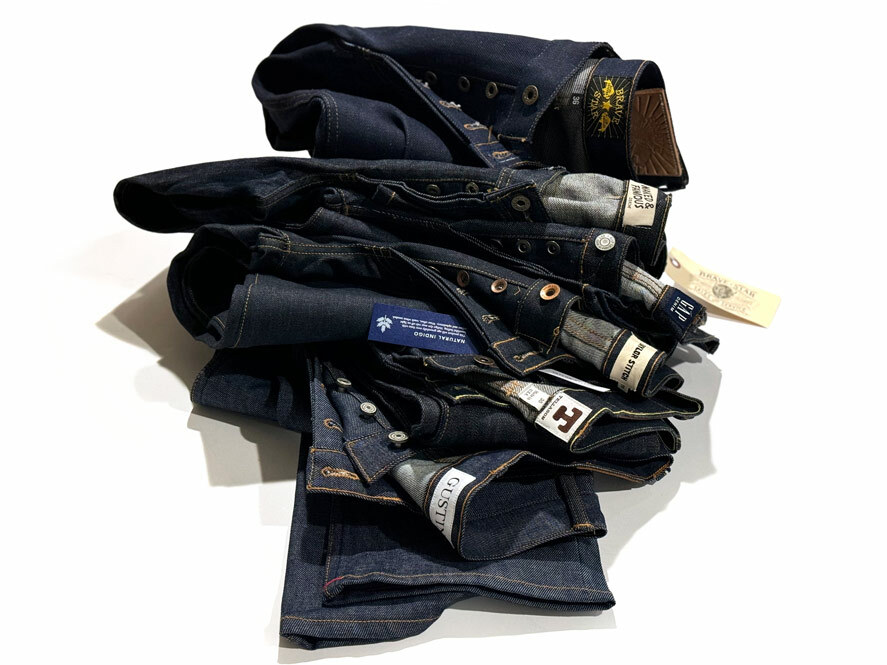 A stack of jeans against a white background, reflecting the numerous raw denim brands that clients send to Williamsburg Garment Company for expert alterations to make their jeans fit perfectly. From top to bottom, this stack features jeans by Brave Star Selvage, Naked and Famous, Gap, Taylor Stitch, Tellason, and Gustin.