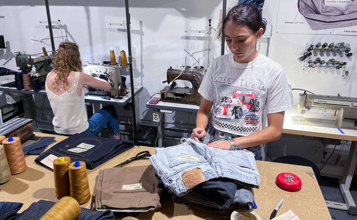 A photograph depicts Williamsburg Garment Company staffers and interns repairing and hemming jeans. Union Special sewing machines and a Singer sewing machine is in the background.