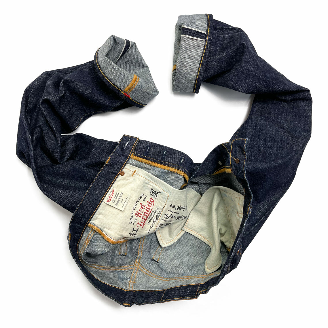 A pair of Red Tornado selvedge denim jeans with chain stitch hemming and a red bartack is artfully arranged to display the front details. The jeans are laid flat, showcasing the rich indigo hue of the raw denim, contrasted by the lighter interior fabric peeking through the unzipped fly. Subtle orange stitching highlights the construction, and a white fabric tag inscribed with care instructions is visible inside the waistband.