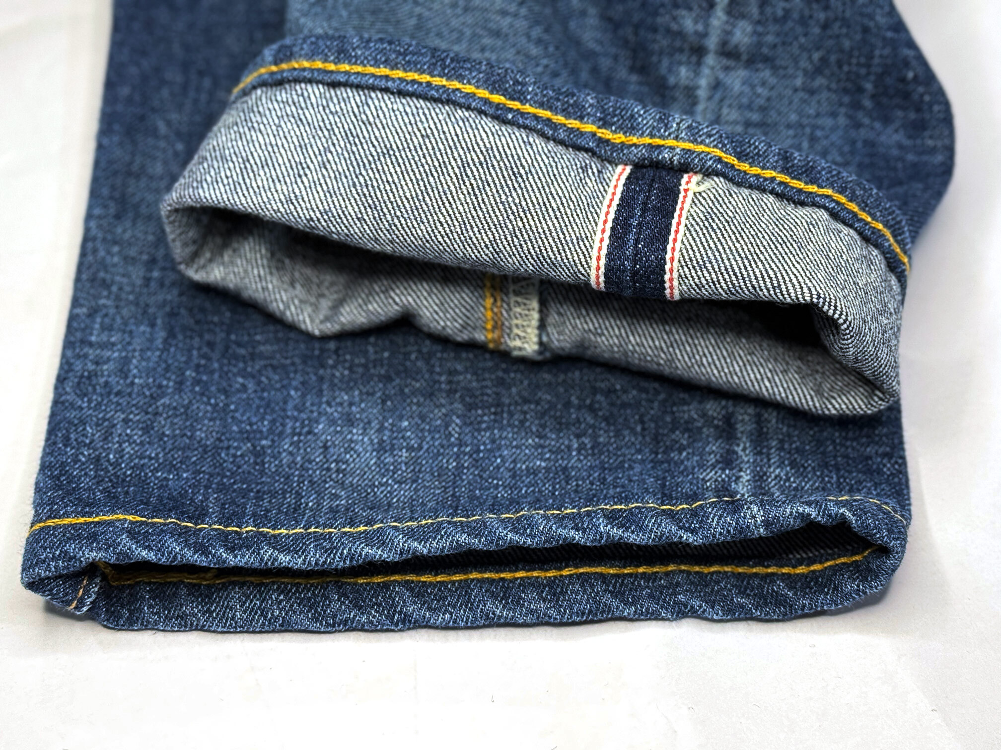 The image depicts a close-up of the hem of selvedge denim jeans, showcasing the chain-stitch sewing and the natural roping effect along the hem edge, which developed after the jeans' first wash.