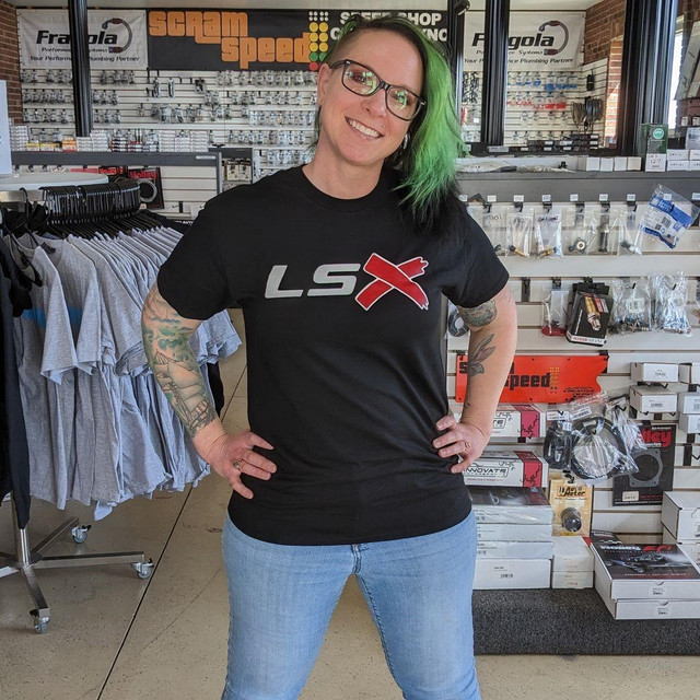 Scram Speed LSX T-shirts, a woman with green and black hair wearing a black 'LSX' logo t-shirt standing in an automotive parts store. Scram Speed LSX T-shirts, a woman with green and black hair wearing a black 'LSX' logo t-shirt standing in an automotive parts store.