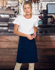 A woman wearing a navy canvas short waist apron over a white t-shirt, standing in a café setting.