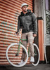 A man wearing cuffed beige stretch pants and a grey jacket stands beside a green bicycle against a textured orange backdrop.
