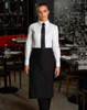 A woman in a black short waist apron over a white shirt and tie, standing in a restaurant setting.