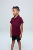A maroon children's polo shirt with a collar, displayed on a young boy wearing black shorts. The shirt has a logo.