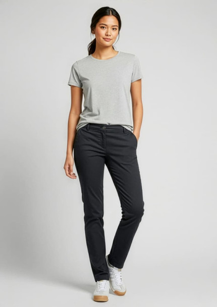 A woman stands in black chino pants and a grey t-shirt, smiling with one hand in her pocket. The image includes a logo.