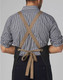A denim bib apron with a cross-back design and a brown tie, worn by a person in a checked shirt.